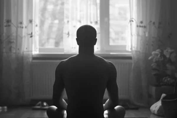 Homem sentado de costas em postura de pernas cruzadas diante de uma janela, exemplificando o que é meditação.